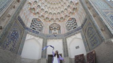 Uzun saçlı, doğulu cüppeli ve kafatası şapkalı genç bir kadın antik bir doğu şehrinde yürüyor. Yavaş Hareket, Özbekistan