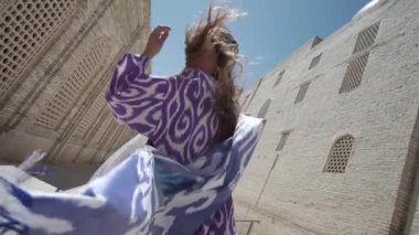 Uzun saçlı, doğulu cüppeli ve kafatası şapkalı genç bir kadın antik bir doğu şehrinde yürüyor. Yavaş Hareket, Özbekistan