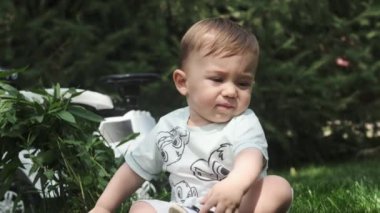 Şortlu küçük bir çocuk parkta çimlerin üzerinde oturur ve plastik bir topla oynar. Yavaş Hareket.