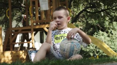 Şortlu, tişörtlü ve çıplak ayaklı bir genç koltuk altında futbol topu ve elinde yeşil bir elmayla parkta çimlerin üzerinde oturuyor. Yavaş Hareket.