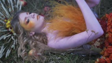 Çiçeklerden, yapraklardan ve dikenlerden oluşan güz tasarımcı kıyafetli genç bir kadın dağınık elmaların arasında çimlerin üzerinde yatıyor. Yavaş Hareket, Kapat.