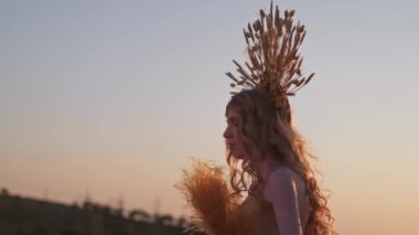 Dikenli başlıklı ve sonbahar çiçekli elbisesi içinde genç bir kadın, elinde yapraklarla elinde kuru dallarla dağlardaki günbatımının arka planında duruyor. Yavaş Hareket