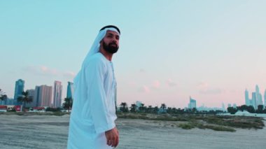 Ulusal Arap elbiseli genç sakallı bir işadamı Dubai 'nin gökdelenlerinin önünde duruyor. Bulutlu bir gökyüzünde gün batımı.