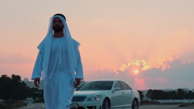 Ulusal Arap elbiseli genç sakallı bir işadamı pahalı arabasından kameraya doğru yürür. Arka planda gün batımı bulutlu gökyüzü.