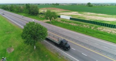Bir İHA otoyolda giden bir kamyonun üzerinden uçuyor. Yolun etrafında yeşil tarlalar ve çayırlar var. Bulutlu bir yaz günü. Hava Görünümü.