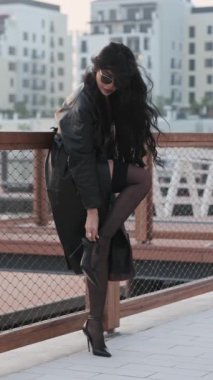 Genç, uzun saçlı esmer, yüksek topuklu ayakkabı ayarlıyor. O, uzun bir pelerin ve çorap giydi. Yavaş Hareket