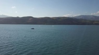 Bir dron dağların arasında gölün üzerinde uçuyor. Bir tekne ve jet ski suda yüzüyor. Güneşli bir yaz günü. Hava Görünümü.