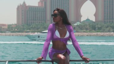 Beyaz bikinili esmer bir kadın ve şehrin arka planına karşı özel bir teknede açık renk bir elbise giyiyor. Ağır çekim, BAE