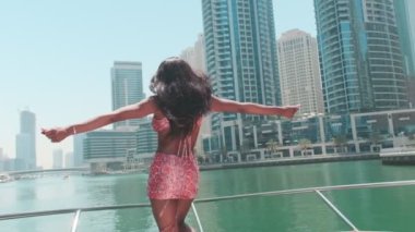 Dubai gökdelenlerinin arka planına karşı özel bir teknenin güvertesinde kısa parlak elbiseli esmer bir kadın. BAE, Ağır çekim