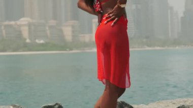 Bikinili esmer ve açık kırmızı elbiseli genç bir kadın denizin ve Dubai 'nin gökdelenlerinin önünde çıplak ayakla kayalarda yürüyor. BAE, Ağır çekim.