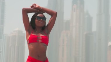 Bikinili esmer ve açık kırmızı elbiseli genç bir kadın denizin ve Dubai 'nin gökdelenlerinin önünde çıplak ayakla kayalarda yürüyor. BAE, Ağır çekim.