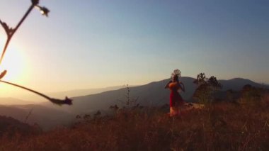 Drone, sonbahar kıyafetiyle günbatımı arka planında dağlardaki çayırlarda genç bir kadının üzerinden uçuyor. Hava görünümü.