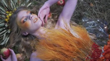 Çiçeklerden, yapraklardan ve dikenlerden oluşan güz tasarımcı kıyafetli genç bir kadın dağınık elmaların arasında çimlerin üzerinde yatıyor. Yavaş Hareket, Kapat.