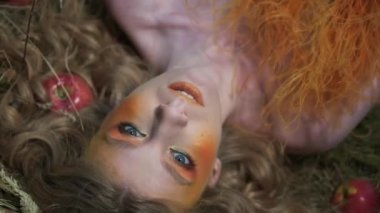Çiçeklerden, yapraklardan ve dikenlerden oluşan güz tasarımcı kıyafetli genç bir kadın dağınık elmaların arasında çimlerin üzerinde yatıyor. Yavaş Hareket, Kapat.