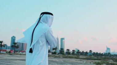 Ulusal Arap elbiseli genç sakallı bir işadamı Dubai 'nin gökdelenlerinin önünde duruyor. Bulutlu bir gökyüzünde gün batımı.