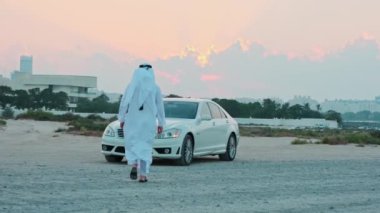 Ulusal Arap elbiseli genç sakallı bir işadamı pahalı arabasına doğru yürür. Dubai 'nin gökdelenlerinin arkasında gün batımı.
