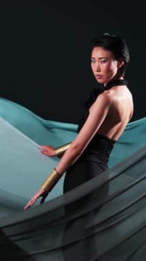 Siyah tulumlu ve parlak makyajlı Asyalı bir gencin dikey stüdyo portresi. Rüzgarda çırpınan bir kumaş