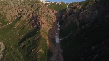 Bir dron buzla dağ geçidinde uçuyor. Etrafta ağaçlar ve çalılar var. Güneşli bir bahar günü. Hava Görünümü
