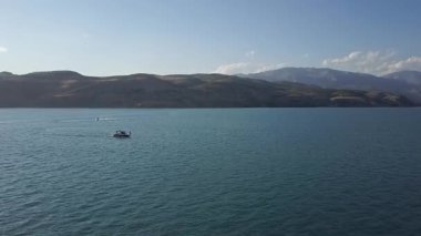 Bir dron dağların arasında gölün üzerinde uçuyor. Bir tekne ve jet ski suda yüzüyor. Güneşli bir yaz günü. Hava Görünümü.