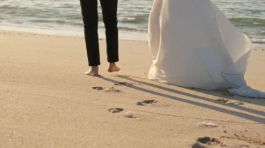 Birbirine aşık genç bir çift kumlu deniz kıyısında yalınayak yürüyor. Güneşli bir yaz günü, balayı. Yavaş Hareket.