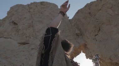 Genç sakallı bir şaman çölün kum tepelerinin arasında bir kaya mağarasında dururken dua eder. Yavaş çekim