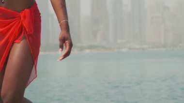 Bikinili esmer ve açık kırmızı elbiseli genç bir kadın denizin ve Dubai 'nin gökdelenlerinin önünde çıplak ayakla kayalarda yürüyor. BAE, Ağır çekim.