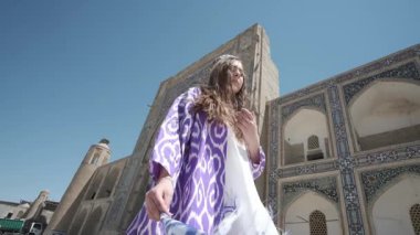 Uzun saçlı, doğulu cüppeli ve kafatası şapkalı genç bir kadın antik bir doğu şehrinde yürüyor. Yavaş Hareket, Özbekistan