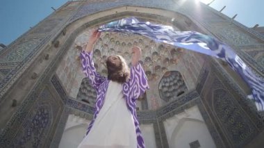 Doğulu cüppeli ve şapkalı, uzun saçlı genç bir kadın eski bir caminin yanında rüzgârda duruyor. Yavaş Hareket, Özbekistan
