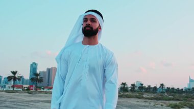 Ulusal Arap elbiseli genç sakallı bir işadamı Dubai 'nin gökdelenlerinin önünde duruyor. Bulutlu bir gökyüzünde gün batımı.