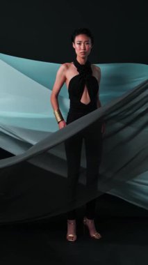 Siyah tulumlu ve parlak makyajlı Asyalı bir gencin dikey stüdyo portresi. Rüzgarda çırpınan bir kumaş