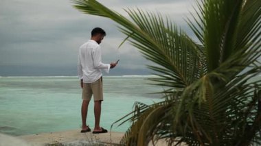 Elinde telefonlar olan açık renk elbiseli genç bir adam palmiye ağaçlarının altında kumlu bir sahilde duruyor. Deniz dalgaları ve bulutlu gökyüzünün arka planına karşı. Maldivler. Yavaş Hareket.