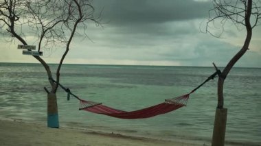 Arka planda deniz olan kumlu bir sahilde, iki ağacın arasına gerilmiş hamak. Sörf dalgaları sahili yıkıyor. Bulutlu bir gün Maldivler. Yavaş Hareket