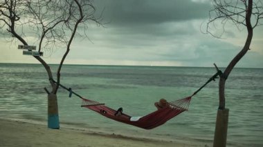 Açık renk elbiseli genç bir adam okyanustaki ağaçların arasında bir hamakta yatıyor. Kumsal, deniz, arka planda bulutlu gökyüzü. Yavaş Hareket