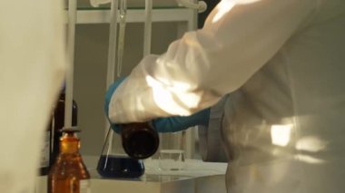 Beyaz önlüklü ve koruyucu eldivenli bir laboratuvar teknisyeni kimyasal bir kaba sıvı döker. Laboratuvar, ağır çekim..