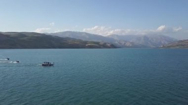 Bir dron dağların arasında gölün üzerinde uçuyor. Bir tekne ve jet ski suda yüzüyor. Güneşli bir yaz günü. Hava Görünümü.