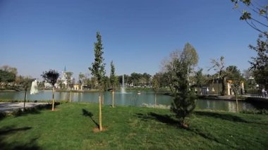 Şehir gölünün kıyısında bir yürüyüş, yeşillik ve ağaçlarla ekilmiş. Gölde fıskiyeler var. Güneşli bir yaz günü. Yavaş Hareket