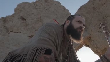 Genç sakallı bir şaman çölün kum tepelerinin arasında bir kaya mağarasında dururken dua eder. Yavaş çekim