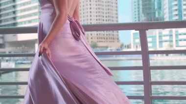Sarışın ve uzun mor elbiseli genç bir kadın marina boyunca Dubais gökdelenlerinin önünde yürüyor. Yavaş Hareket.