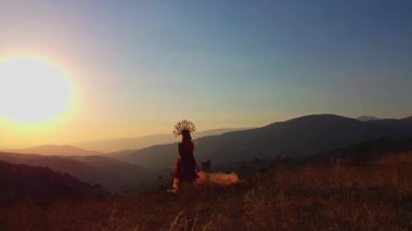 Drone, sonbahar kıyafetiyle günbatımı arka planında dağlardaki çayırlarda genç bir kadının üzerinden uçuyor. Hava görünümü.