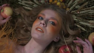 Çiçeklerden, yapraklardan ve dikenlerden oluşan güz tasarımcı kıyafetli genç bir kadın dağınık elmaların arasında çimlerin üzerinde yatıyor. Yavaş Hareket, Kapat.