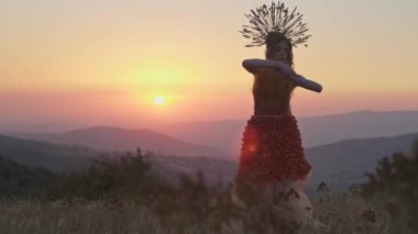 Dikenli başlıklı genç bayan manken ve dağlarda günbatımının arka planında çiçeklerden ve otlardan yapılmış elbise. Odaklanma ön plandan arka plana hareket eder. Yavaş Hareket.