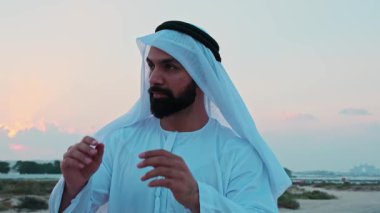 Ulusal Arap elbiseli genç sakallı bir işadamı Dubai 'nin gökdelenlerinin önünde duruyor. Bulutlu bir gökyüzünde gün batımı.