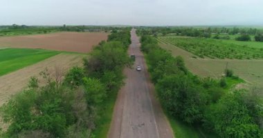 Bir İHA, kır yolunda giden bir kamyonun üzerinden uçuyor. Etrafta ağaçlar ve tarlalar var. Bulutlu bir yaz günü. Hava Görünümü.