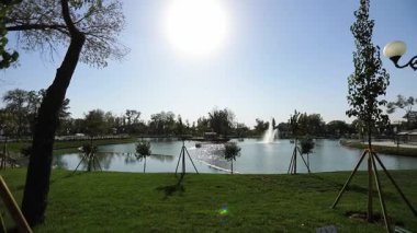 Şehir gölünün kıyısında bir yürüyüş, yeşillik ve ağaçlarla ekilmiş. Gölde fıskiyeler var. Güneşli bir yaz günü. Yavaş Hareket