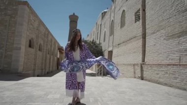 Uzun saçlı, doğulu cüppeli ve kafatası şapkalı genç bir kadın antik bir doğu şehrinde yürüyor. Yavaş Hareket, Özbekistan