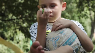 Şortlu, tişörtlü ve çıplak ayaklı bir genç koltuk altında futbol topu ve elinde yeşil bir elmayla parkta çimlerin üzerinde oturuyor. Yavaş Hareket, Kapat.