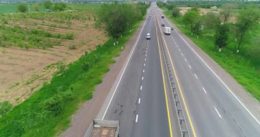 Bir İHA otobanda giden bir kamyonun önünde uçuyor. Etrafta hareket eden arabalar, yeşil tarlaların arasında bir yol var. Bulutlu bir yaz günü. Hava Görünümü.