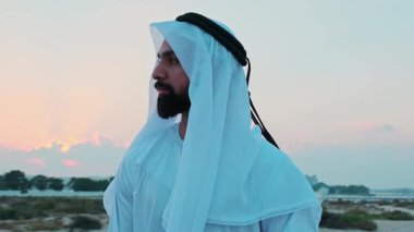 Ulusal Arap elbiseli genç sakallı bir işadamı Dubai 'nin gökdelenlerinin önünde duruyor. Bulutlu bir gökyüzünde gün batımı.