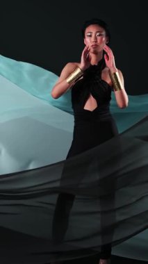 Siyah tulumlu ve parlak makyajlı Asyalı bir gencin dikey stüdyo portresi. Rüzgarda çırpınan bir kumaş