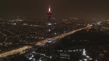 Parlak bir televizyon kulesiyle aydınlık bir şehrin üzerinde bir dron. Bulutsuz bir yaz gecesi. Hava Görünümü.